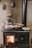 A traditional wood-burning stove with a visible fire inside the stove door. Several aluminum trays with cooked food and a shiny metal kettle are placed on top of the stove. The background features a textured wall adorned with vintage, decorative copper molds.