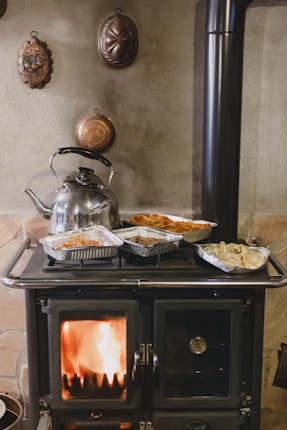 A traditional wood-burning stove with a visible fire inside the stove door. Several aluminum trays with cooked food and a shiny metal kettle are placed on top of the stove. The background features a textured wall adorned with vintage, decorative copper molds.