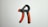 A hand gripper with an adjustable spring is placed on a plain white surface. The handles are black with textured orange grip sections.