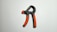 A hand gripper with an adjustable spring is placed on a plain white surface. The handles are black with textured orange grip sections.