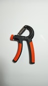 A handheld grip strengthener with black and orange handles is positioned against a plain white background. The design features a spring mechanism at the top, allowing for adjustable resistance.