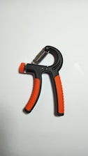 A handheld grip strengthener with black and orange handles is positioned against a plain white background. The design features a spring mechanism at the top, allowing for adjustable resistance.