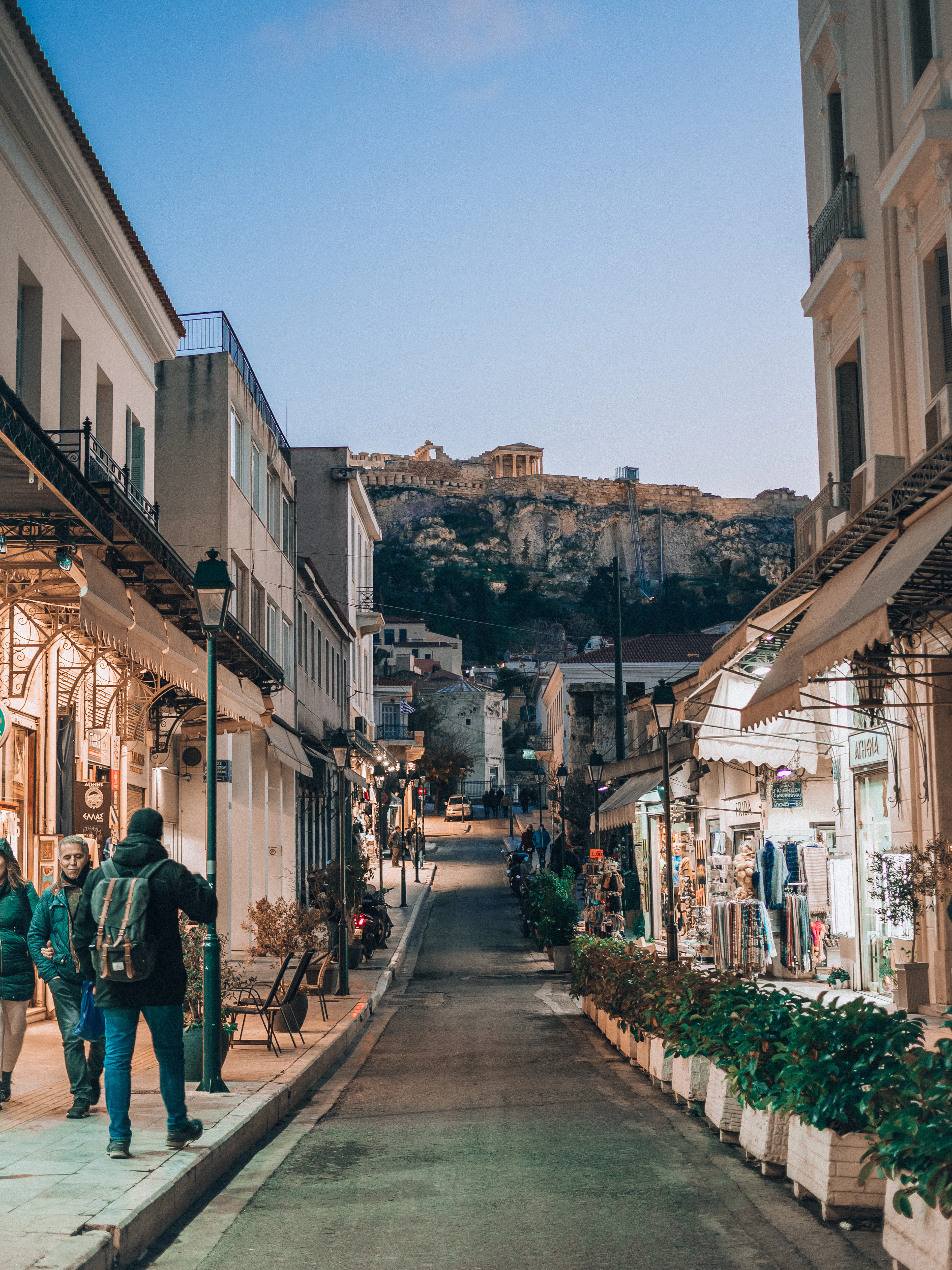 A shopping street in Plaka, Athens, at dusk, neoclassical balconies and shop awnings running down both sides and the Parthenon standing on the Acropolis rock straight ahead at the end of the road