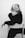 A person with a short, tousled hairstyle is seated on a chair, wearing a dark outfit. The image is in black and white, creating a stark contrast between the light and dark tones. The person is looking directly at the camera with a contemplative expression, one hand resting on their head.