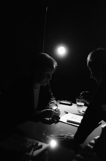 A dark, mysterious setting with two individuals engaged in conversation under a single overhead light. Shadows obscure much of their features, creating a moody atmosphere. A desk is cluttered with papers and a glass of water.