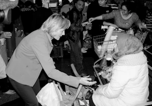A woman in a warm coat and headscarf is seated in front of a shopping cart filled with various items. She is engaged in conversation with a standing woman, who gestures with her hand towards the seated woman. Several other people are present in the background, some holding items and appearing to distribute goods.