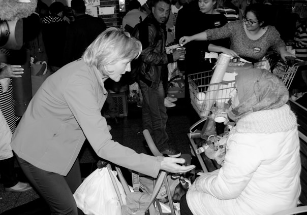 A woman in a warm coat and headscarf is seated in front of a shopping cart filled with various items. She is engaged in conversation with a standing woman, who gestures with her hand towards the seated woman. Several other people are present in the background, some holding items and appearing to distribute goods.