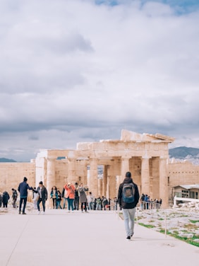 A group of tourists are gathered near the ancient ruins of a temple with large columns. The sky is overcast with clouds, and a person is walking towards the ruins wearing a backpack. The scene conveys a sense of exploration and historical significance.
