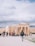 A group of tourists are gathered near the ancient ruins of a temple with large columns. The sky is overcast with clouds, and a person is walking towards the ruins wearing a backpack. The scene conveys a sense of exploration and historical significance.