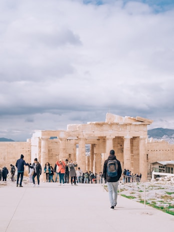 A group of tourists are gathered near the ancient ruins of a temple with large columns. The sky is overcast with clouds, and a person is walking towards the ruins wearing a backpack. The scene conveys a sense of exploration and historical significance.