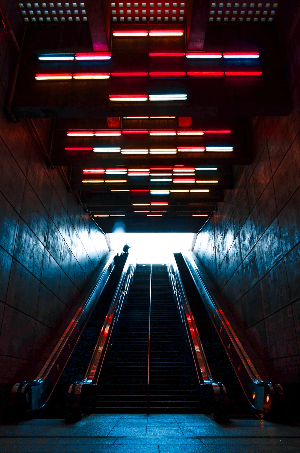 Escalator with red, blue, and green lights illuminates subway station stairs and platform below