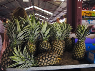 A collection of ripe pineapples arranged on a market stall, with some boxes visible in the background and a few people walking in the marketplace.
