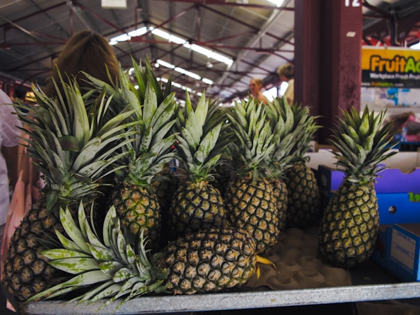 A collection of ripe pineapples arranged on a market stall, with some boxes visible in the background and a few people walking in the marketplace.