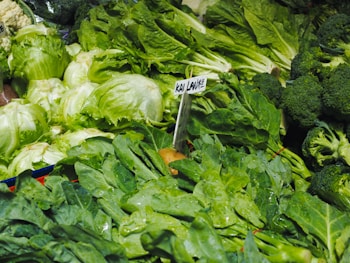 An assortment of fresh, vibrant green vegetables displayed in a market setting. The image features a variety of leafy greens such as lettuce, kai lan (Chinese kale), and broccoli, with some cauliflower visible in the background. The vegetables appear fresh and are likely covered with a light mist of water droplets.