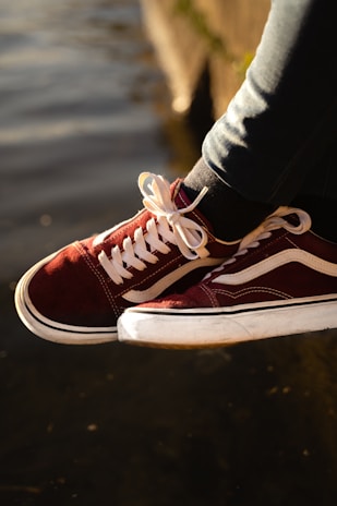 A pair of maroon and white sneakers with white laces is worn by a person sitting near a body of water. The sneakers are illuminated by soft, warm sunlight, creating a relaxed and casual atmosphere.