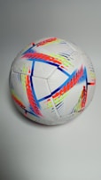 A soccer ball with a vibrant, multicolored pattern featuring shades of red, blue, yellow, and orange on a white background. The design includes geometric shapes and zigzag lines, appearing dynamic and modern against a plain, neutral backdrop.