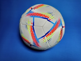 A vibrant soccer ball featuring a complex pattern of colorful, abstract designs in red, blue, neon yellow, and black against a white background, set on a blue backdrop.