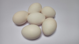 Six light-colored eggs are grouped together on a plain, smooth surface. The arrangement highlights their smooth, oval shapes and their uniform color against the white backdrop.