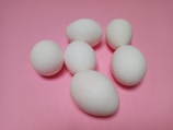 Six white eggs are scattered on a smooth, pastel pink surface. The eggs vary in orientation and are arranged in an informal cluster.