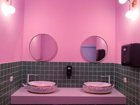 A stylish bathroom features pink walls and a green tiled backsplash. Two round mirrors hang above the sinks, which are placed on a minimalist counter. The sinks have a decorative floral design. Modern light fixtures with spherical bulbs are mounted on the walls, and a soap dispenser and paper towel holder are visible.