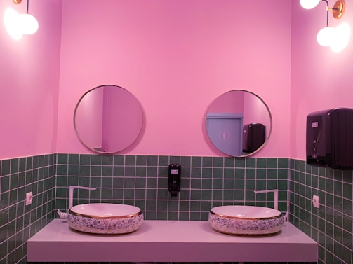 A stylish bathroom features pink walls and a green tiled backsplash. Two round mirrors hang above the sinks, which are placed on a minimalist counter. The sinks have a decorative floral design. Modern light fixtures with spherical bulbs are mounted on the walls, and a soap dispenser and paper towel holder are visible.