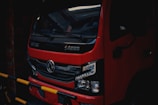 A red truck is parked in a dimly lit area, with a black grille and detailed headlights visible at the front. The vehicle has Chinese characters on the front and a logo in the center. Yellow and black striped barriers are partially obstructing the view.