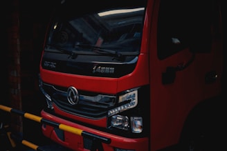 A red truck is parked in a dimly lit area, with a black grille and detailed headlights visible at the front. The vehicle has Chinese characters on the front and a logo in the center. Yellow and black striped barriers are partially obstructing the view.