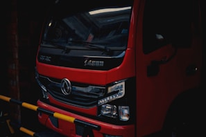 A red truck is parked in a dimly lit area, with a black grille and detailed headlights visible at the front. The vehicle has Chinese characters on the front and a logo in the center. Yellow and black striped barriers are partially obstructing the view.
