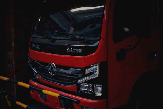 A red truck is parked in a dimly lit area, with a black grille and detailed headlights visible at the front. The vehicle has Chinese characters on the front and a logo in the center. Yellow and black striped barriers are partially obstructing the view.