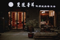 A warmly lit storefront with signs in traditional Chinese characters above the entrance and on the glass doors. The entrance is flanked by potted plants, and inside, tables and chairs are visible, suggesting a cozy tea shop or cafe setting. The interior is softly illuminated with warm lighting, adding to the inviting atmosphere.