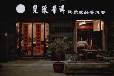 A warmly lit storefront with signs in traditional Chinese characters above the entrance and on the glass doors. The entrance is flanked by potted plants, and inside, tables and chairs are visible, suggesting a cozy tea shop or cafe setting. The interior is softly illuminated with warm lighting, adding to the inviting atmosphere.