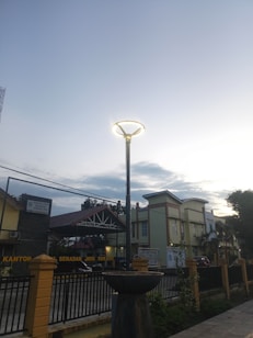 A streetlight with a glowing circular top stands tall against a backdrop of a dusky sky. Below, there is a building with signage and a gated fence, indicative of an official or governmental establishment. The surroundings include some trees and a paved pathway, suggesting an urban environment nearing twilight.