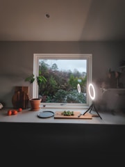 A cozy kitchen scene with a window overlooking a lush garden. A countertop is adorned with a cutting board and knife, as well as a few Brussels sprouts. A round ring light illuminates the area. On the left, a potted plant and some wooden cutting boards are visible, alongside a few oranges.