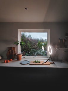 A cozy kitchen scene with a window overlooking a lush garden. A countertop is adorned with a cutting board and knife, as well as a few Brussels sprouts. A round ring light illuminates the area. On the left, a potted plant and some wooden cutting boards are visible, alongside a few oranges.