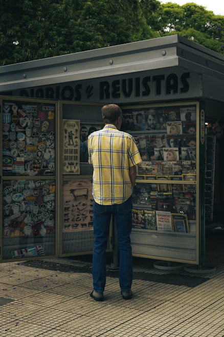 A person wearing a yellow plaid shirt and blue jeans is standing in front of a newsstand filled with various magazines, newspapers, and stickers on display. The newsstand has a sign reading 'DIARIOS y REVISTAS'. Green trees can be seen in the background.