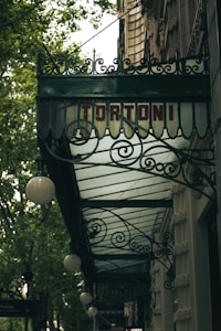 An ornate metal awning with intricate scrollwork is attached to a building. The word 'TORTONI' is prominently displayed in red letters. Street lamps are suspended from the awning, and leafy trees are visible, indicating a street view.