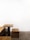 A minimalist wooden table and bench set against a stark white wall. The furniture features clean lines and a simple design, and the bench is positioned at a right angle to the table. The floor is a textured gray concrete, complementing the modern aesthetic.