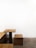 A minimalist wooden table and bench set against a stark white wall. The furniture features clean lines and a simple design, and the bench is positioned at a right angle to the table. The floor is a textured gray concrete, complementing the modern aesthetic.