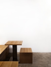 A minimalist wooden table and bench set against a stark white wall. The furniture features clean lines and a simple design, and the bench is positioned at a right angle to the table. The floor is a textured gray concrete, complementing the modern aesthetic.