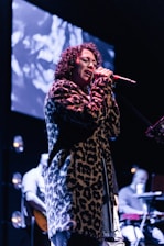 A person with curly hair is passionately singing into a microphone on stage. They are wearing a leopard print coat. The background features a large screen displaying abstract visuals, and there are musicians playing instruments, partially visible, adding to the performance atmosphere.