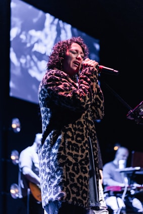 A person with curly hair is passionately singing into a microphone on stage. They are wearing a leopard print coat. The background features a large screen displaying abstract visuals, and there are musicians playing instruments, partially visible, adding to the performance atmosphere.
