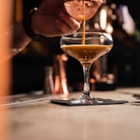 A bartender pouring a rich, dark cocktail into a glass with a cream-colored rim.
