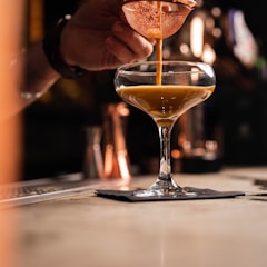 A close-up of a hand pouring a caramel-colored liquid through a strainer into a cocktail glass. The glass rests on a black napkin on a bar counter, with a warm, dimly-lit background suggesting a cozy bar atmosphere.