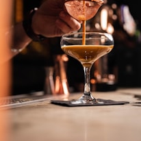 Close-up of a hand pouring mezcal into a rustic glass with soft yellow lighting.