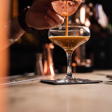 A close-up of a hand pouring a caramel-colored liquid through a strainer into a cocktail glass. The glass rests on a black napkin on a bar counter, with a warm, dimly-lit background suggesting a cozy bar atmosphere.