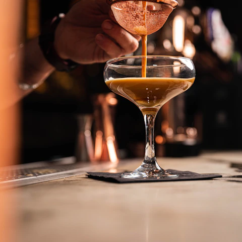 Moody close-up of a hand pouring golden genever into a crystal glass against a dark wood bar backdrop.