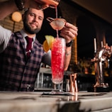 A bartender wearing a plaid vest and red tie is carefully pouring a vibrant red cocktail through a strainer into a tall glass filled with crushed ice, garnished with a slice of orange and lemon. The setting is a stylish bar with sleek counter surfaces and muted lighting, offering a warm, inviting atmosphere.