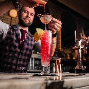 A bartender wearing a plaid vest and red tie is carefully pouring a vibrant red cocktail through a strainer into a tall glass filled with crushed ice, garnished with a slice of orange and lemon. The setting is a stylish bar with sleek counter surfaces and muted lighting, offering a warm, inviting atmosphere.