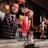 A bartender wearing a plaid vest and red tie is carefully pouring a vibrant red cocktail through a strainer into a tall glass filled with crushed ice, garnished with a slice of orange and lemon. The setting is a stylish bar with sleek counter surfaces and muted lighting, offering a warm, inviting atmosphere.