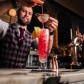 A bartender wearing a plaid vest and red tie is carefully pouring a vibrant red cocktail through a strainer into a tall glass filled with crushed ice, garnished with a slice of orange and lemon. The setting is a stylish bar with sleek counter surfaces and muted lighting, offering a warm, inviting atmosphere.